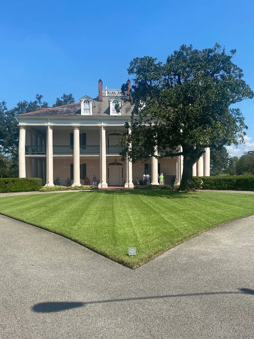The image shows a grand two-story colonial-style house with a columned portico set in manicured grounds under a clear blue sky