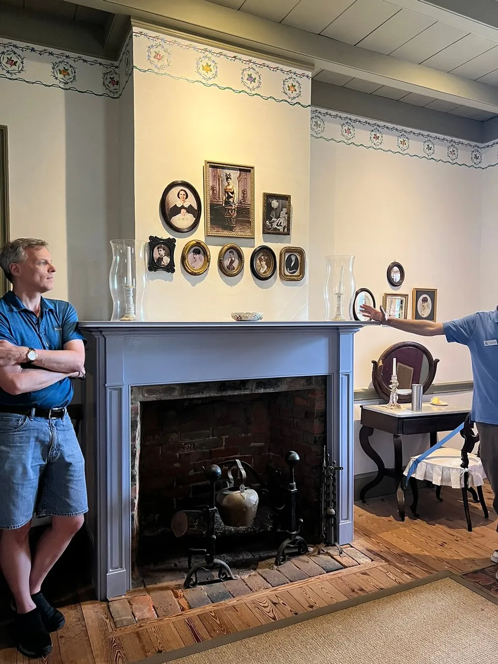 A man stands to the left arms crossed looking at an antique fireplace beneath a decorated wall with framed portraits while another persons arm is visible to the right in a room with vintage furnishings and decor