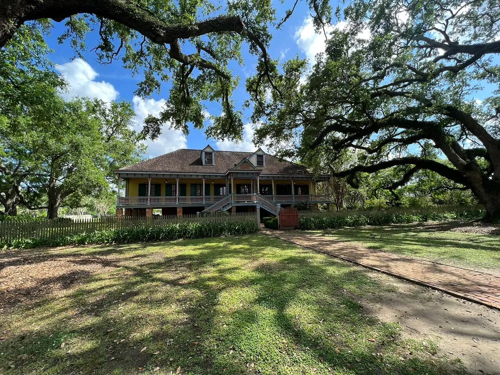 This image showcases a charming two-story house with a large porch nestled among mature oak trees in a grassy landscape