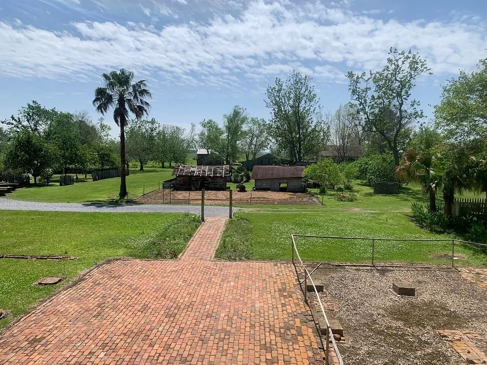 The image depicts a sunny green rural landscape with a brick pathway leading towards a group of rustic wooden buildings and a palm tree suggesting a historical or agricultural setting