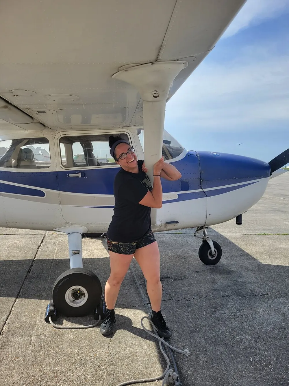 A smiling woman is playfully posing with her arms around the strut of a small single-engine airplane parked on the tarmac