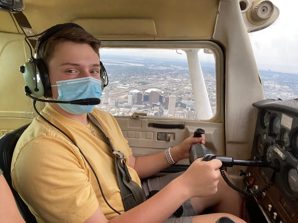 A young person wearing a mask and aviation headset is seated at the controls of a small aircraft with a cityscape visible through the window