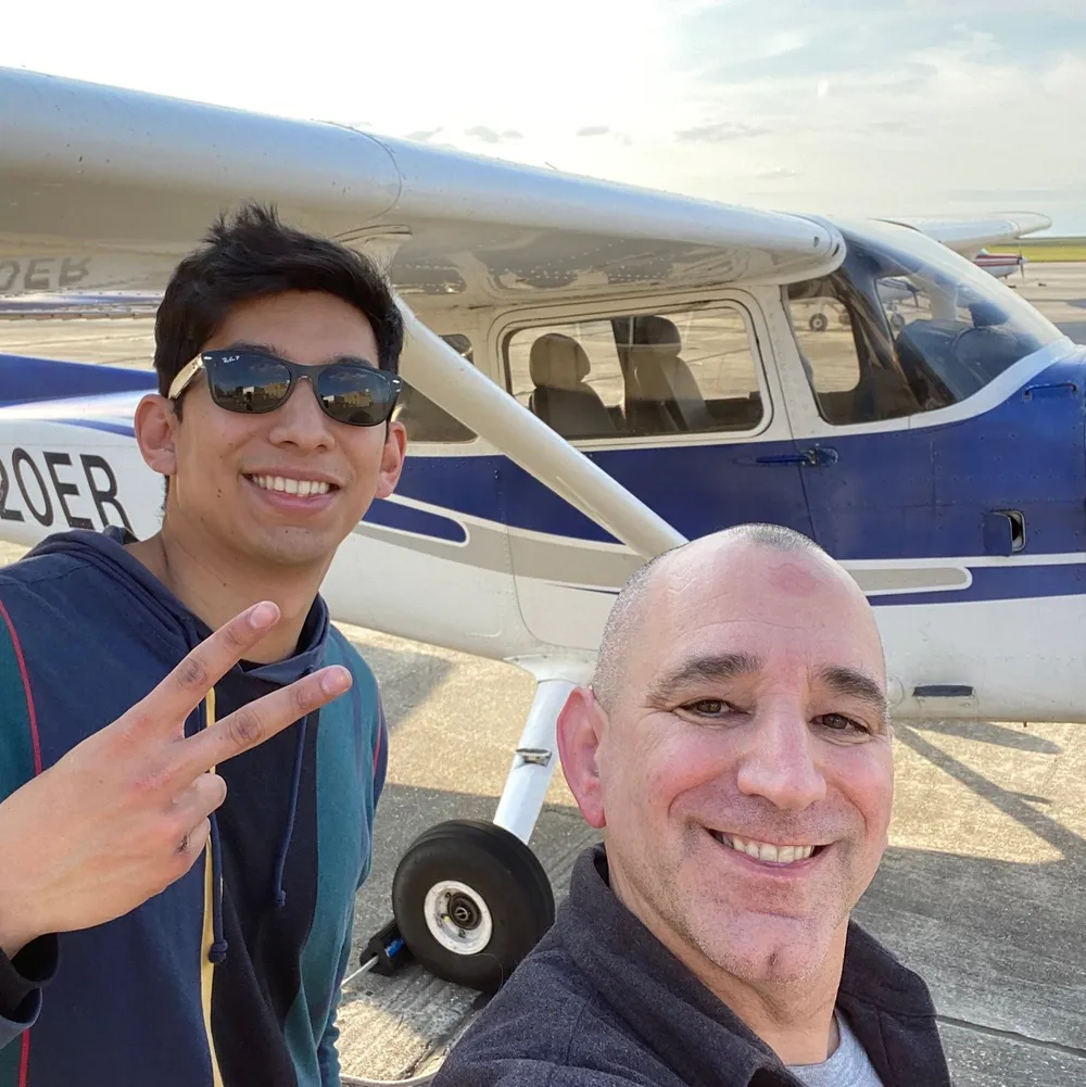 Two smiling individuals are taking a selfie in front of a blue and white single-engine airplane on a sunny day