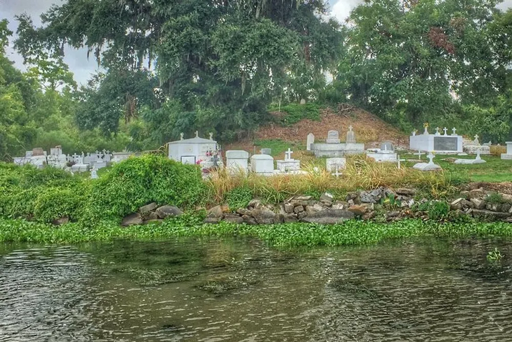 A cemetery with various white tombstones and crosses is situated on a lush green hillside beyond a body of water under a cloudy sky