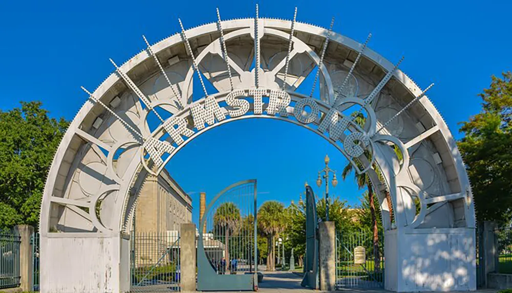 The image shows a large decorative metal archway with the word ARMSTRONG at the top marking the entrance to a park or public space set against a clear blue sky
