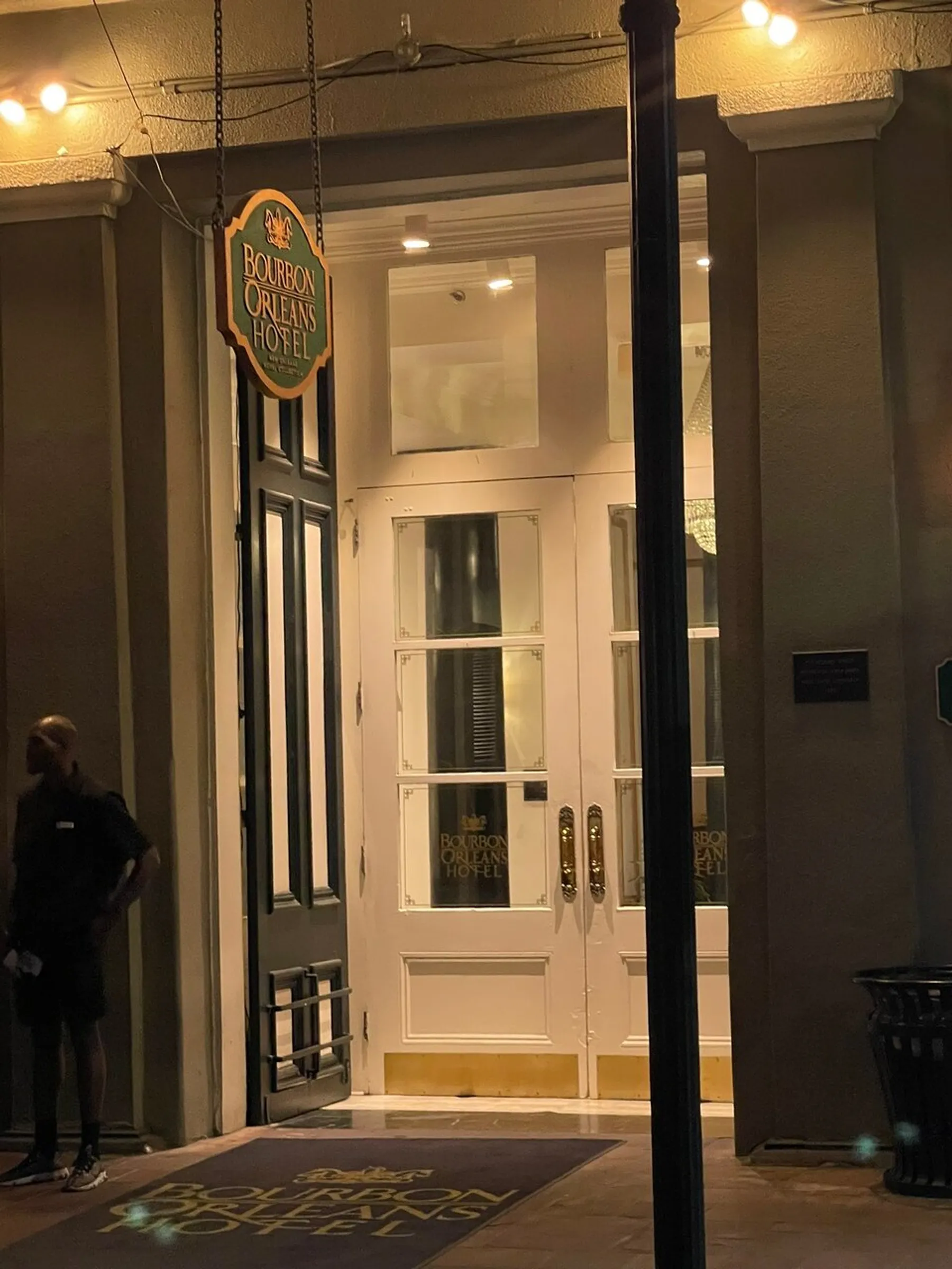 The image shows the illuminated entrance of the Bourbon Orleans Hotel at night, with a person standing by the door and the hotel's ornate sign hanging above.
