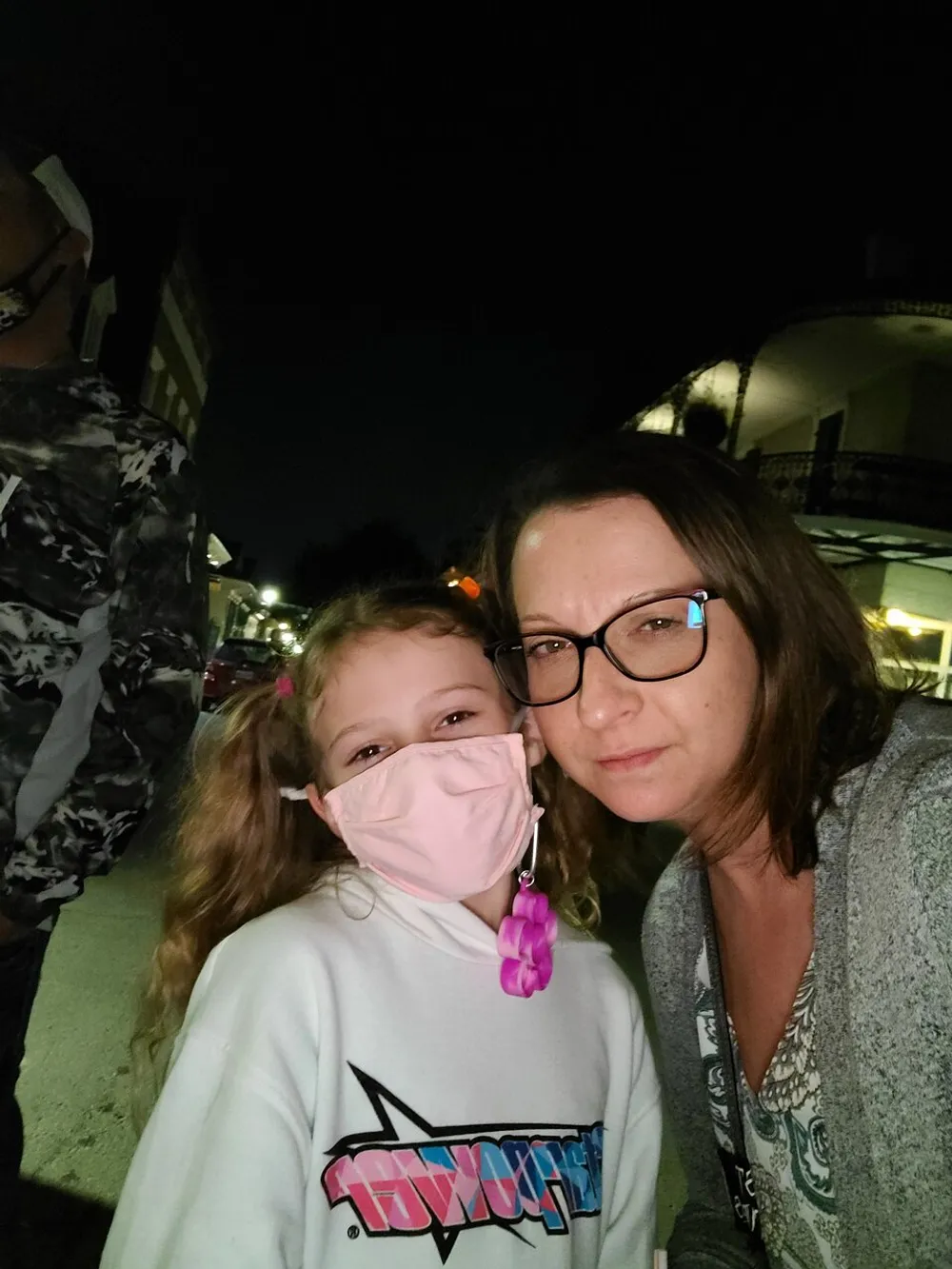 A smiling child wearing a pink face mask and an adult are posing closely together for a selfie at night with the child showing wearable accessories and the adult wearing glasses