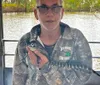 A person is holding a small alligator while wearing a camouflage jacket with Louisiana Tour Company text on it standing inside a boat with a river and trees visible in the background