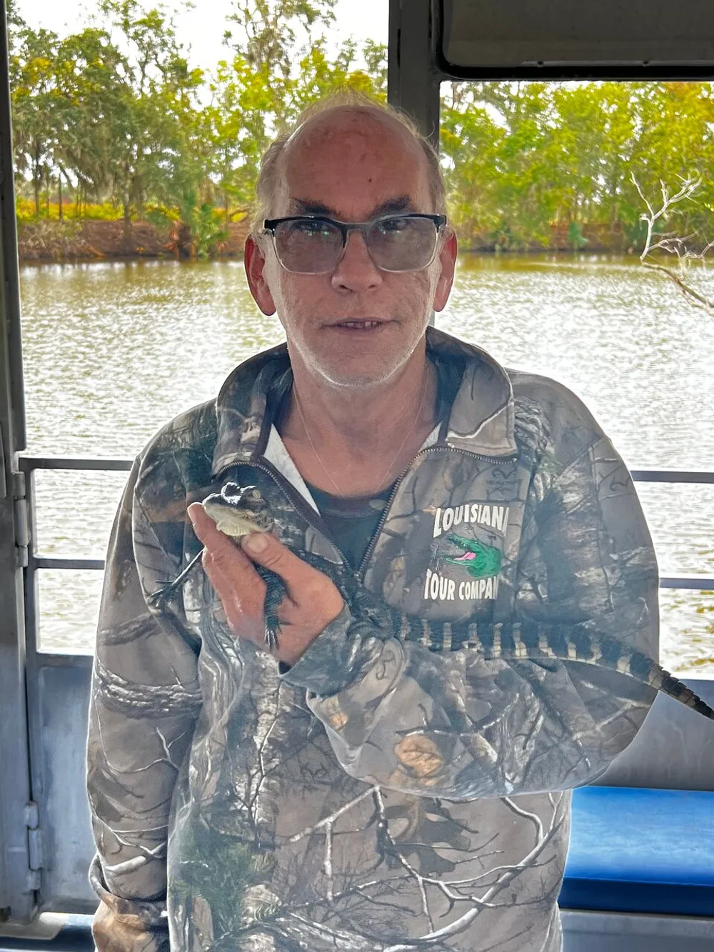 A person is holding a small alligator while wearing a camouflage jacket with Louisiana Tour Company text on it standing inside a boat with a river and trees visible in the background