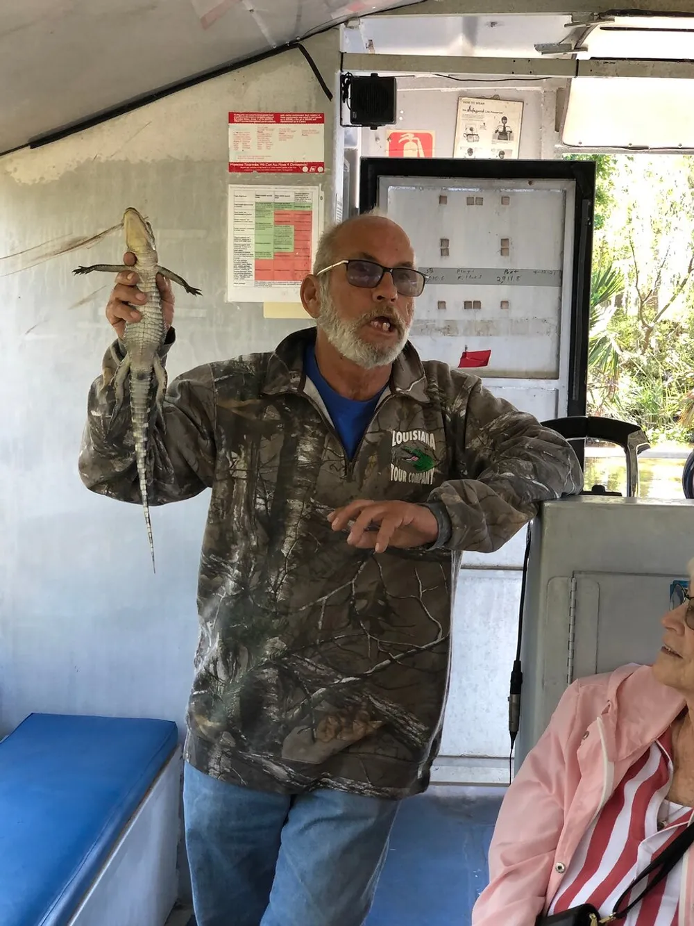 A man is holding a small alligator while giving a presentation on a boat as a passenger looks on