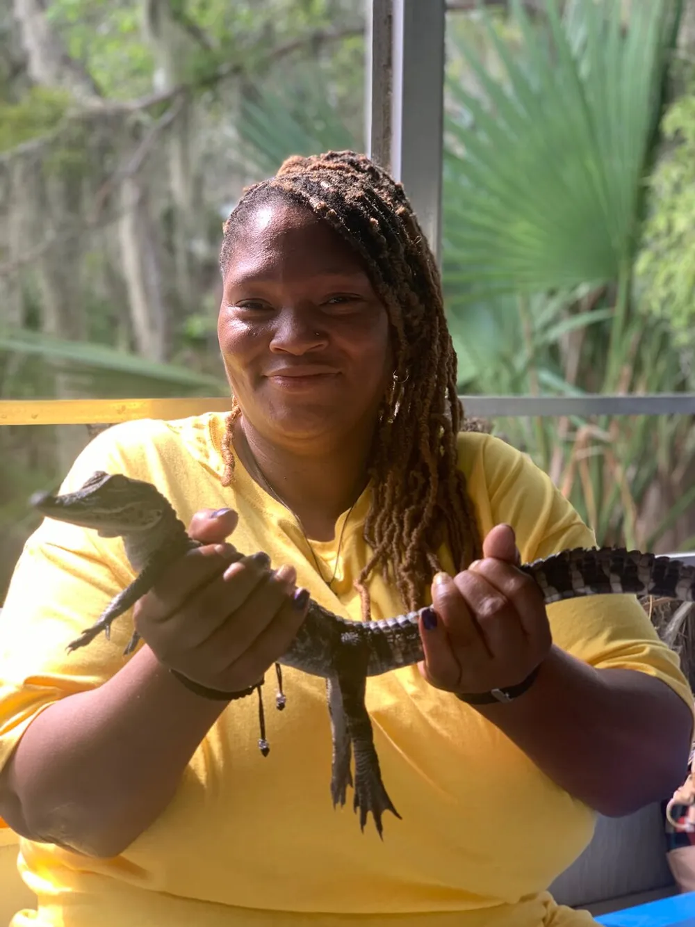 A smiling person is holding a small alligator with a backdrop of greenery