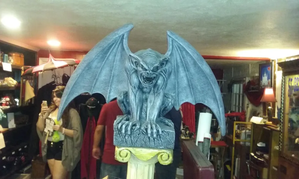 The image shows a large gargoyle statue with outstretched wings atop a pedestal in an indoor setting that appears to be an eclectic shop with various individuals and items in the background