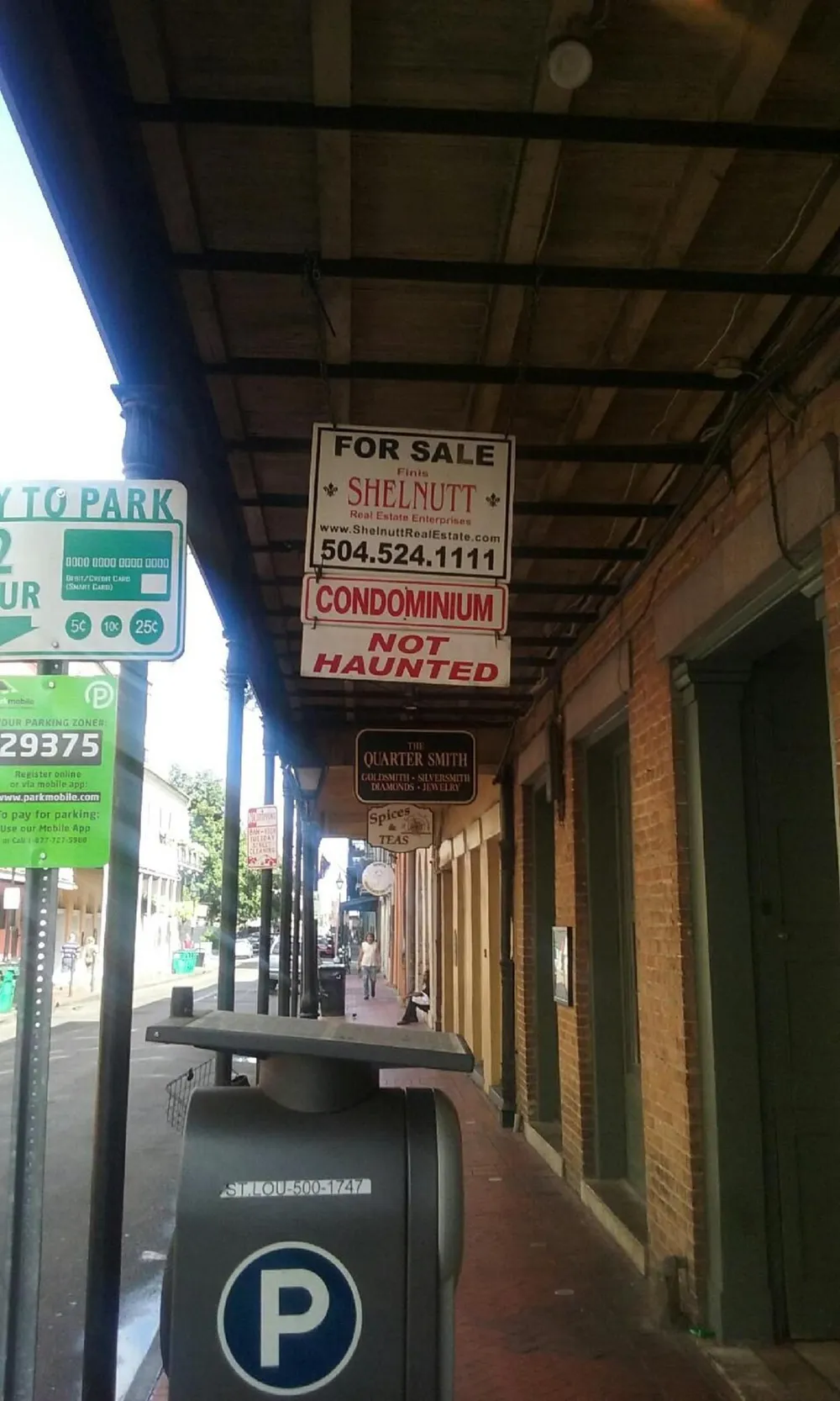The image shows an urban street scene with a For Sale sign for a condominium that humorously notes it is NOT HAUNTED hung above a sidewalk in what appears to be an older neighborhood