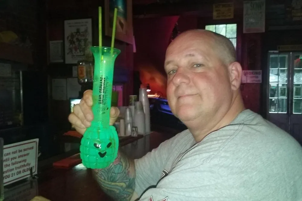 A man is smiling at the camera while holding up a green novelty drink served in a tall skull-shaped glass with two straws at a bar