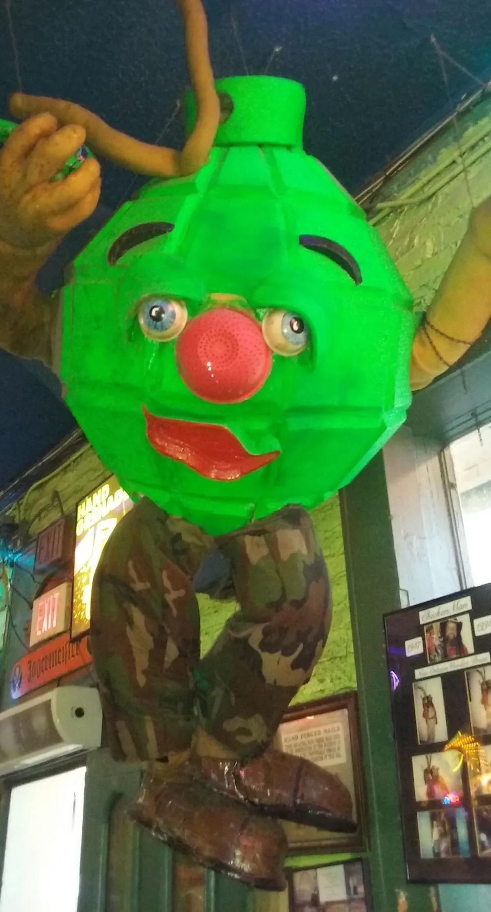 The image features a quirky oversized green character with a red clown nose and camouflage pants hanging from a ceiling being adjusted by a persons hand