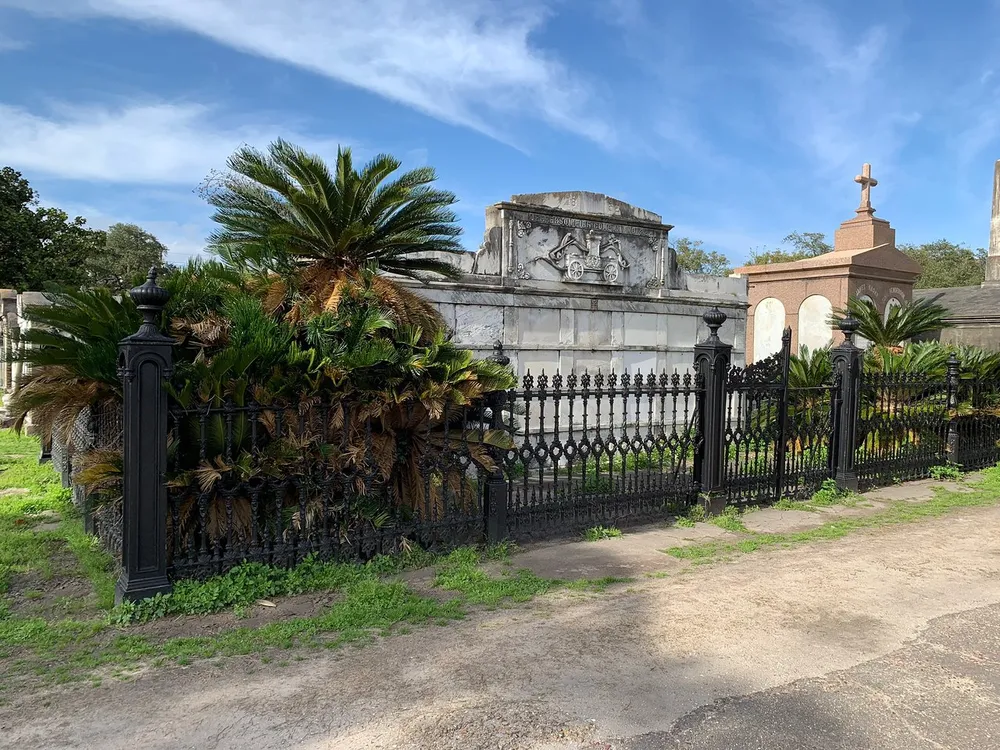 The image shows a sunny view of an ornate historic cemetery with above-ground tombs and iron fencing flanked by lush palm trees