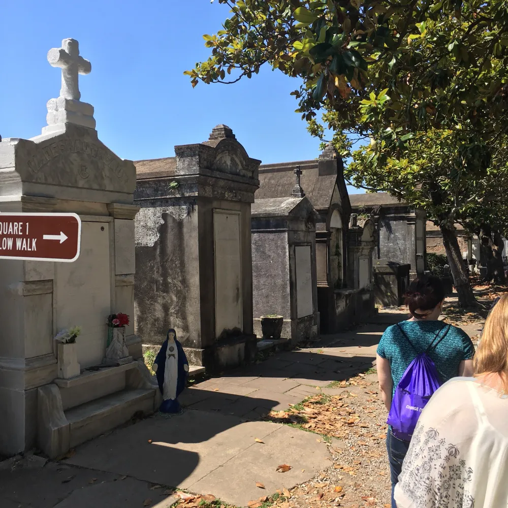 Visitors are walking through a historic cemetery with above-ground tombs and a statue of the Virgin Mary on a sunny day