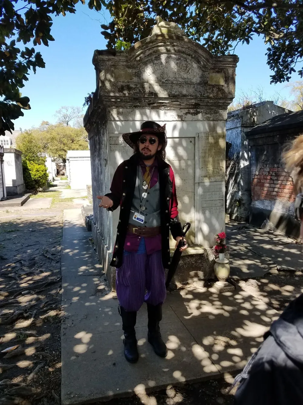 A person dressed in historically-inspired attire stands before an old tombstone in what appears to be a historical or ghost tour