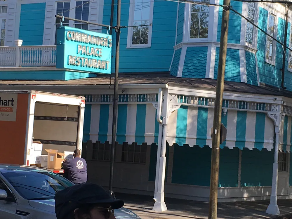 The image shows a vibrant blue building with a sign that reads Commanders Palace Restaurant with an individual and vehicles in the foreground indicating an urban setting