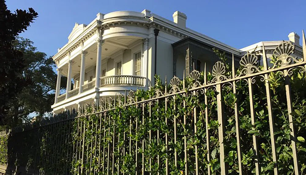 The image shows a grand white house with a wraparound balcony behind an ornate metal fence surrounded by lush greenery under a clear blue sky