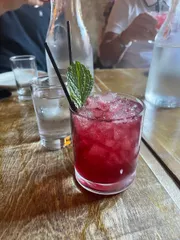 A refreshing crimson-colored cocktail with ice and a mint garnish is served on a wooden table with blurred background of people and other drinks.