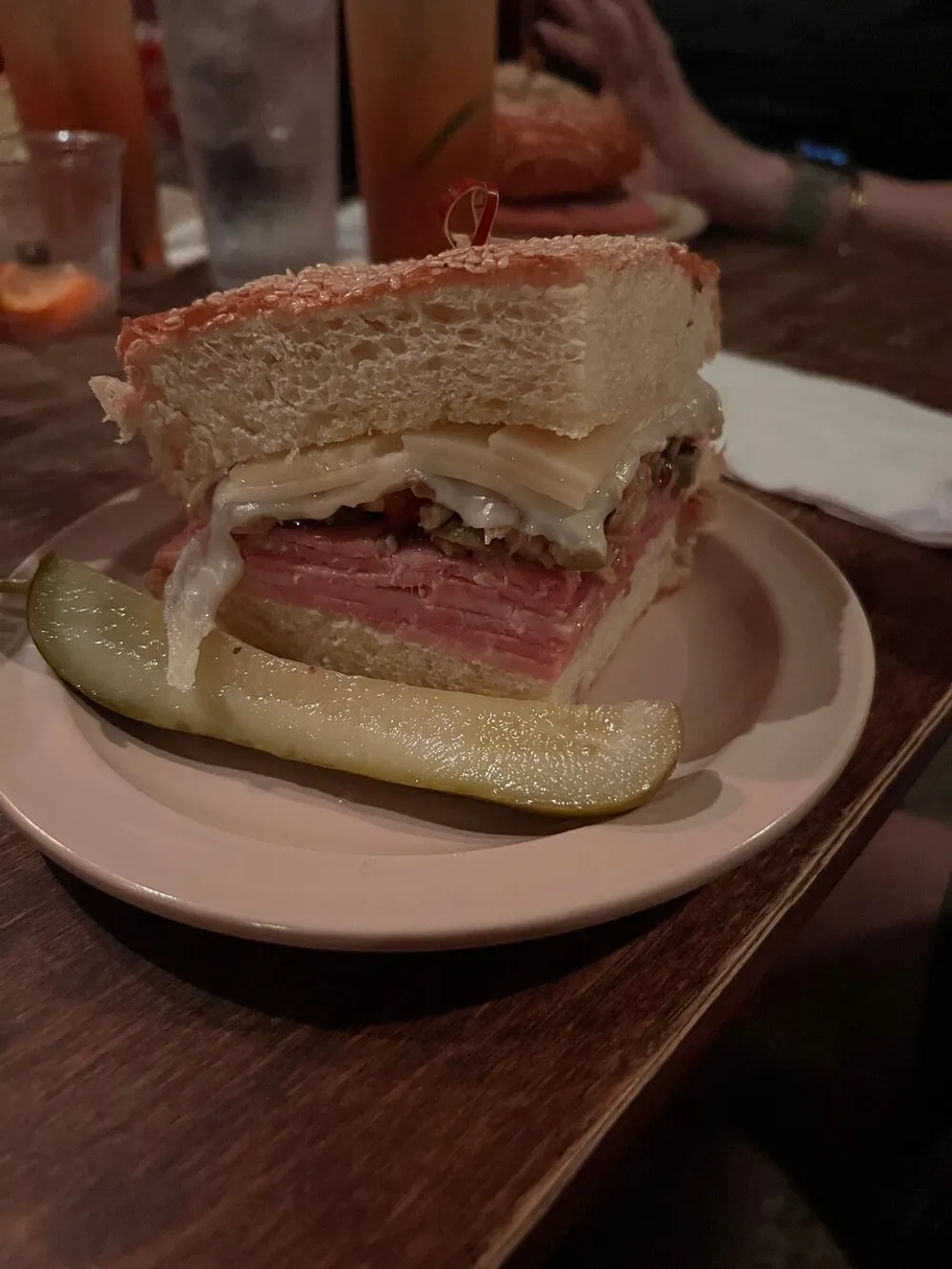 The image showcases a thickly layered sandwich on a plate next to a pickle spear with a blurred background indicating a casual dining setting