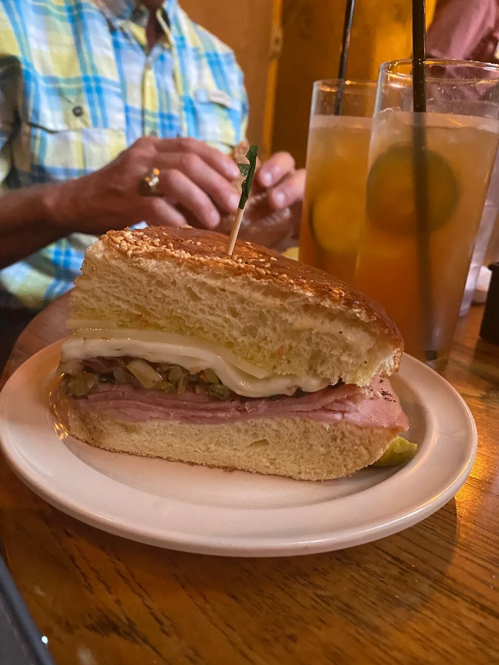 A half-eaten sandwich with ham cheese and pickles is on a plate in the foreground with a person and iced drinks in the background