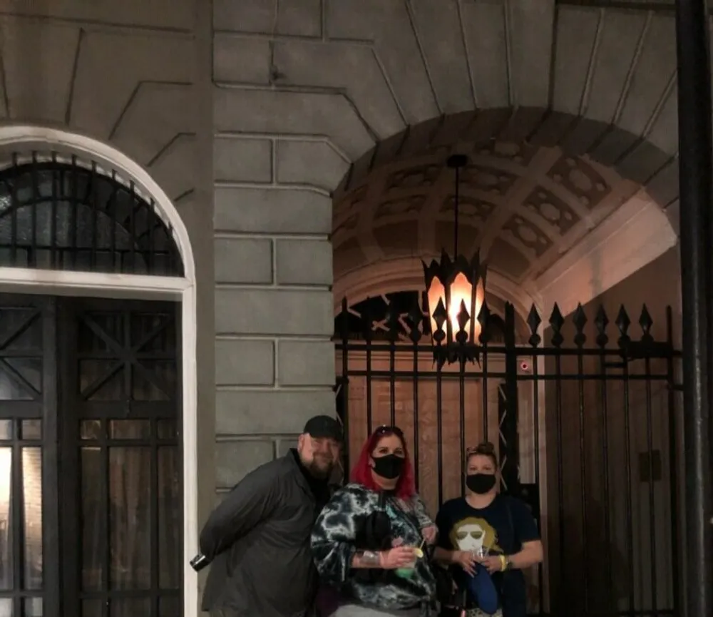 Three individuals are posing for a photo in front of a gated entrance with an ornate lamp fixture above them at night