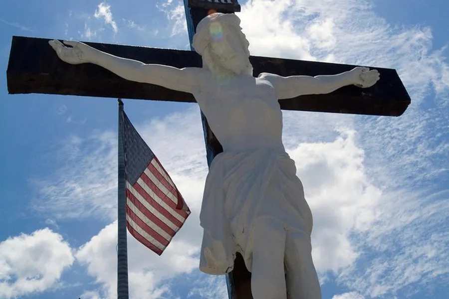 The image shows a statue of Jesus on the cross situated next to an American flag against a backdrop of blue sky and clouds.