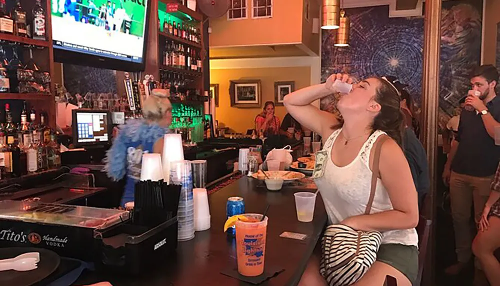 A person is tilting their head back to drink a shot at a lively bar with other patrons and a bartender in the background