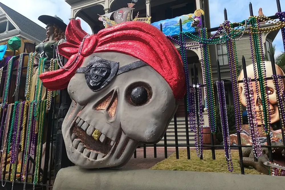 The image features a large skull with glittering red fabric and a pirate symbol on its head set against a backdrop of a decorated house with Mardi Gras beads and other whimsical decorations suggesting a festive pirate-themed celebration