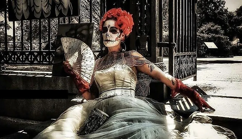 A person is dressed in an elaborate costume with a skull face makeup reminiscent of the Mexican La Catrina sitting on a bench with a fan in hand evoking a scene from the Day of the Dead celebrations