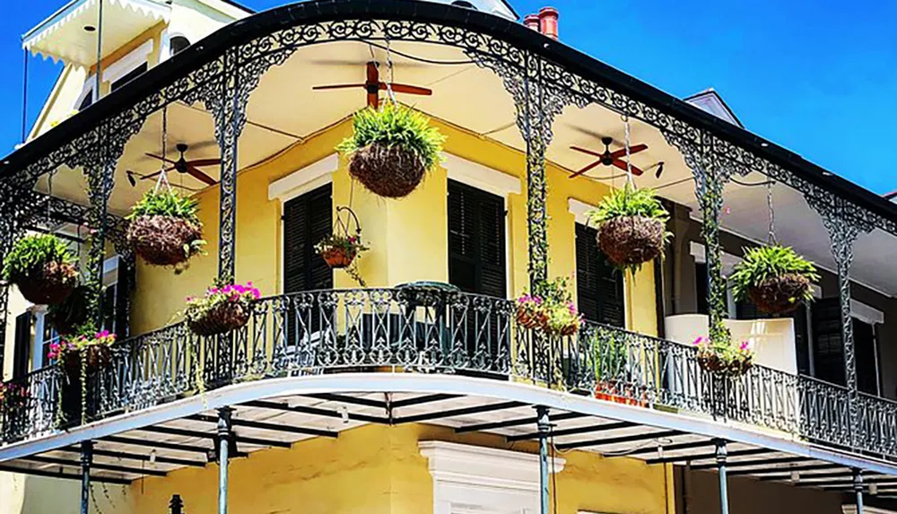 A yellow building with ornate black iron balconies adorned with hanging plants and ceiling fans under a clear blue sky