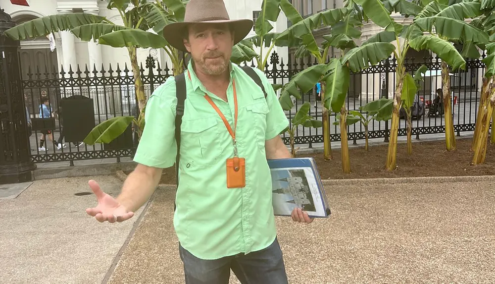 A man wearing a hat and a green shirt is standing outdoors holding a tablet in one hand with an outstretched arm and looking at the camera