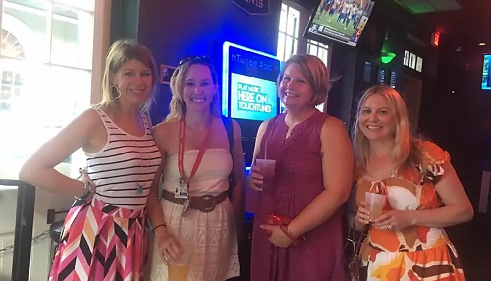 Four women are smiling for a photo inside a bar holding drinks and dressed in casual summer attire