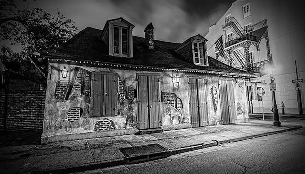 A black and white photograph captures an old weathered building with shutters on the windows situated on a quiet street at night conveying a haunting atmosphere
