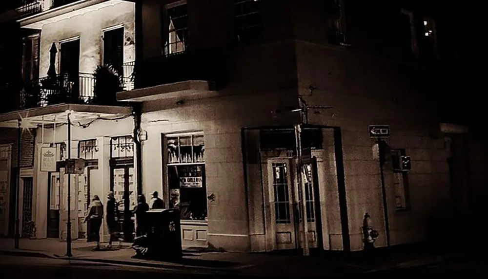 A sepia-toned image captures a quaint and dimly lit street corner at night where people are gathered outside a vintage-looking storefront