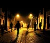 The image captures a dimly lit atmospheric cobblestone street at night with silhouetted figures of people walking and creating a moody and somewhat mysterious scene