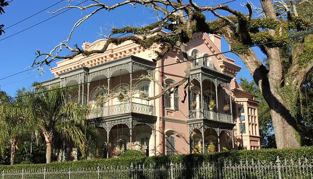 The image shows a two-story pink Victorian house with intricate ironwork on the balconies and a lush green garden partially obscured by a large tree all set against a clear blue sky