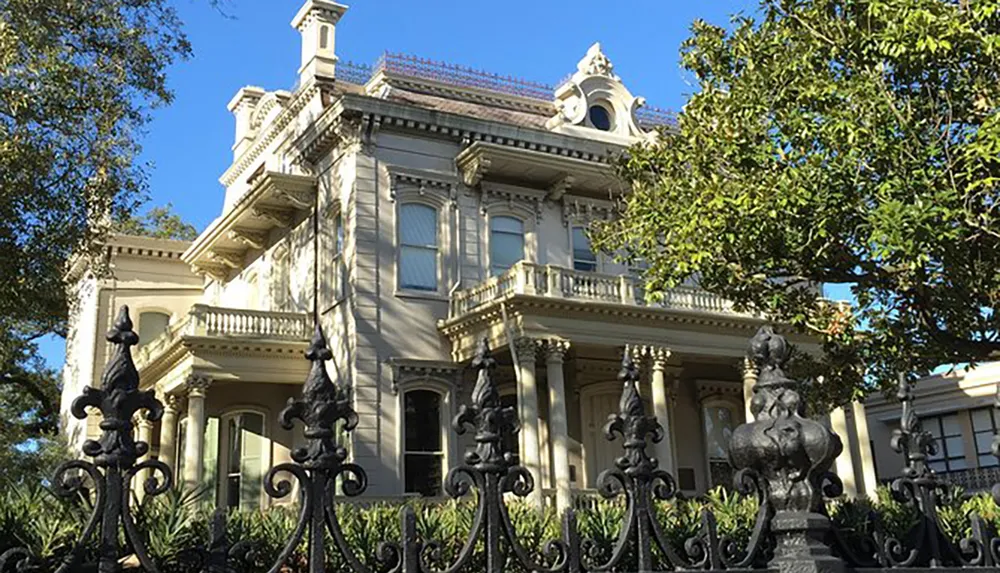 The image shows a grand historic mansion with ornate detailing a balcony and an intricate wrought iron fence set against a backdrop of clear skies and greenery