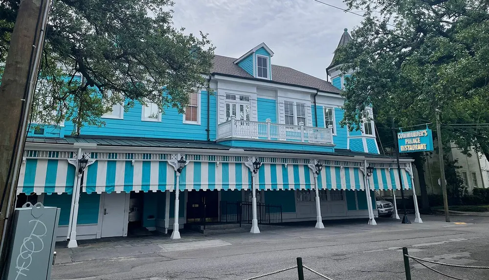 The image shows a vibrant blue two-story building with white and blue striped awnings adorned by street lamps with a sign indicating its Commanders Palace Restaurant
