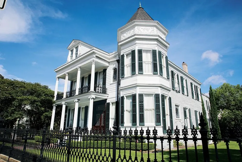 The image shows a grand white Victorian-style house with a prominent tower feature expansive porches black wrought-iron fencing and neatly manicured lawn under a clear blue sky