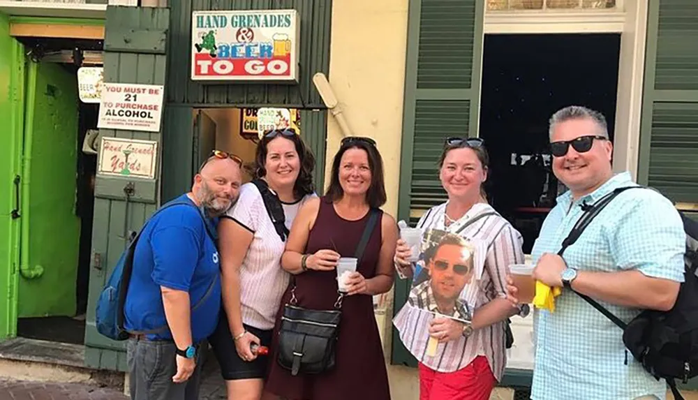 A group of five smiling people some holding beverages pose in front of a store with a sign reading Hand Grenades indicating they might be enjoying a social outing