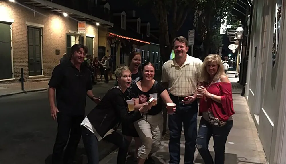 A group of people are smiling and posing for a photo on a sidewalk at night with some holding drinks