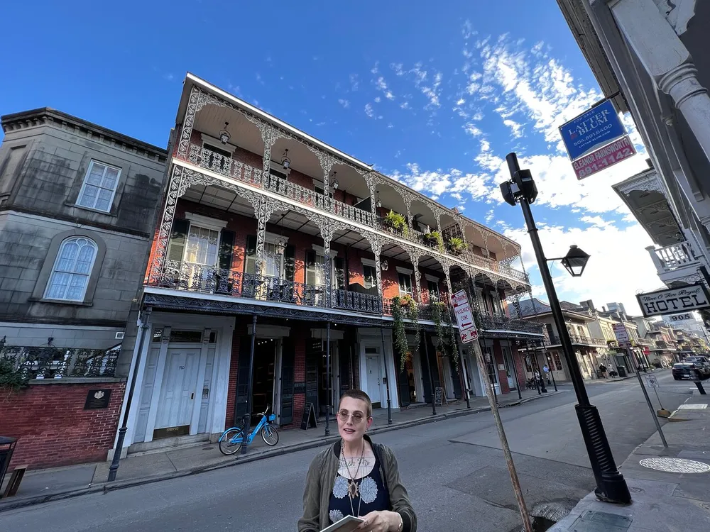 A person stands in a sunny street with traditional wrought-iron balconies in what appears to be the French Quarter of New Orleans