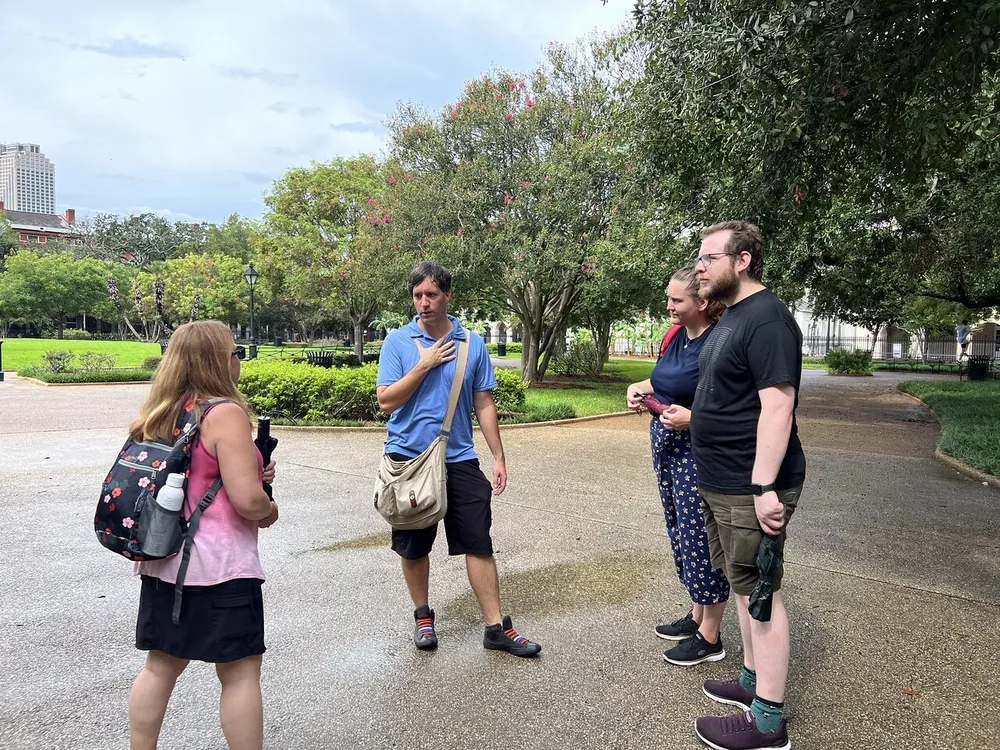A group of people appear engaged in a discussion or a tour outdoors in a park with trees and walkways in the background
