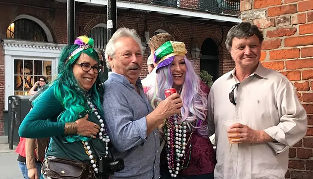 Four people are smiling and posing for a photo at an outdoor event wearing colorful Mardi Gras beads and accessories with two of them holding drinks