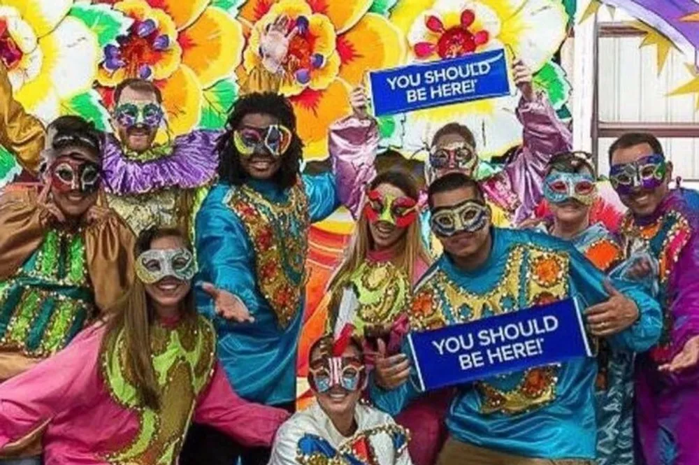 A jubilant group of people in vibrant carnival costumes and masks holding signs that say YOU SHOULD BE HERE are posing together