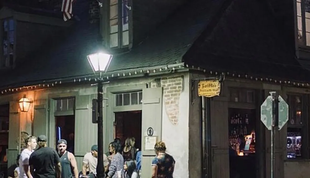 The image shows a group of people standing and chatting outside a cozy corner building at night that appears to be a bar or pub illuminated by street lamps
