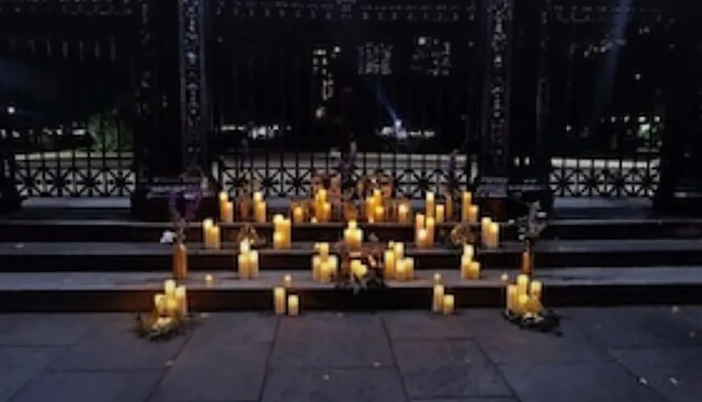 The image shows a serene nighttime vigil with numerous candles lit on some steps likely as a tribute or memorial